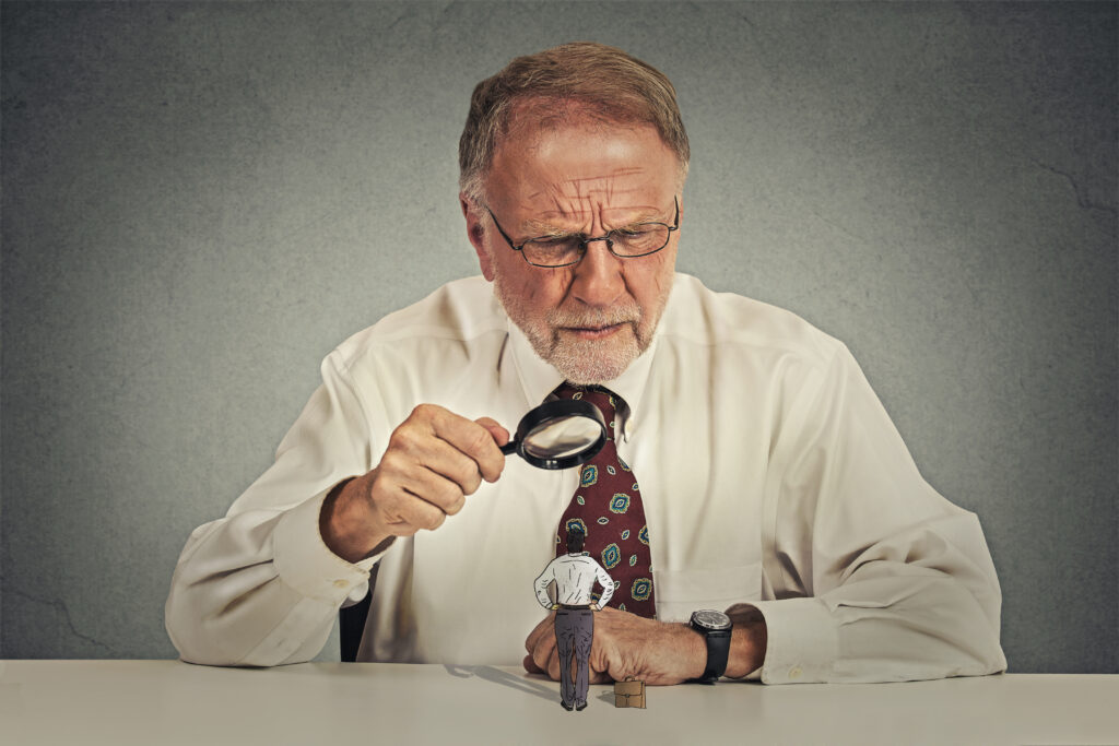Curious corporate senior businessman skeptically meeting looking at small employee worker through magnifying glass isolated office grey wall background. Human face expression, attitude, perception